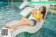 A woman in a yellow bikini sitting on a lounge chair by a pool.