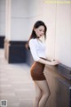 A woman leaning against a wall in a white shirt and brown skirt.