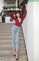 A woman in a red top and jeans leaning against a wall.