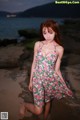 A woman in a floral dress posing on a rock by the water.