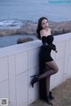 A woman in a black dress leaning against a wall by the ocean.