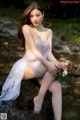 A woman in a white lingerie sitting on a rock by a stream.